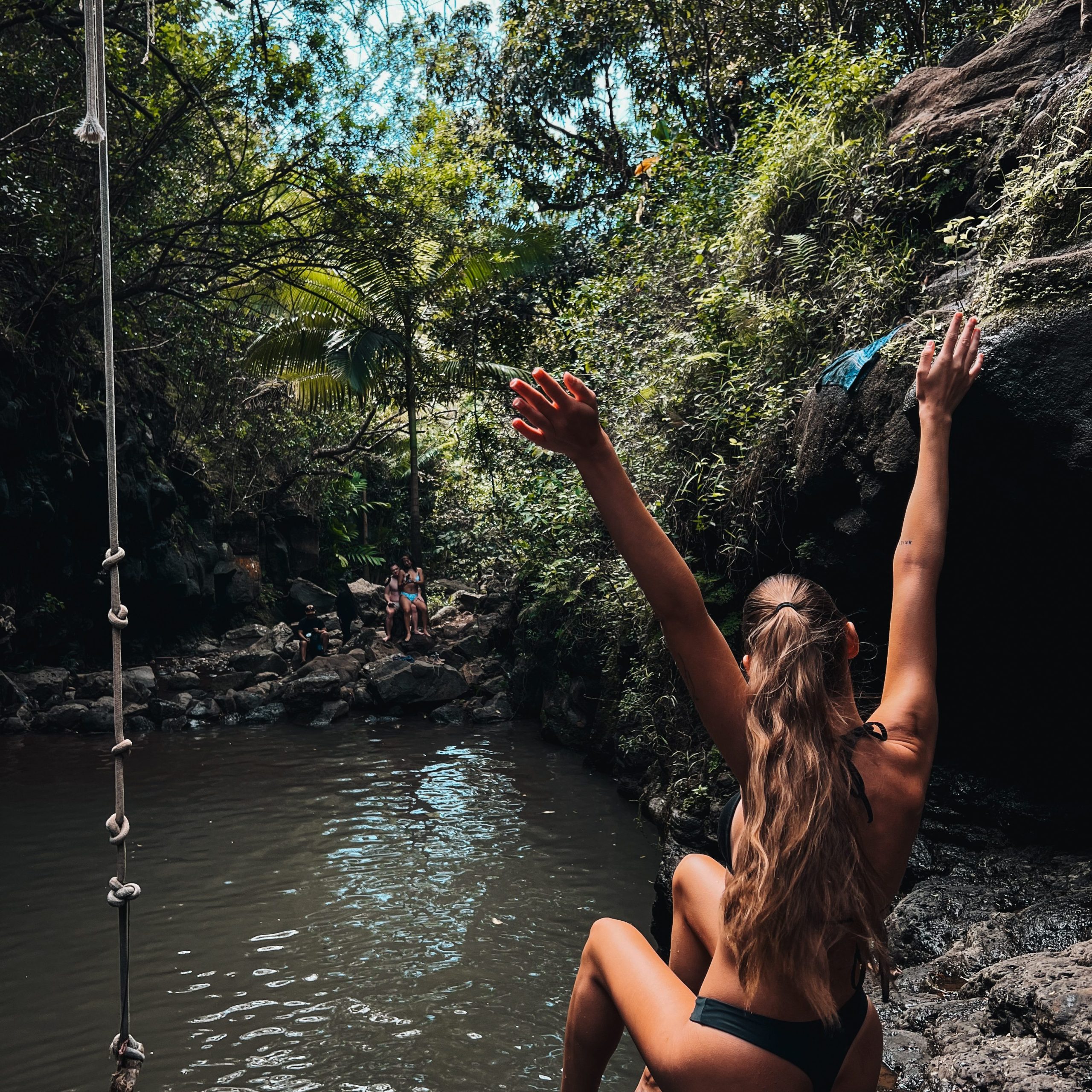 Girl sitting in front of some water in the jungel in Hawaii, being happy having her hands up in the sky.